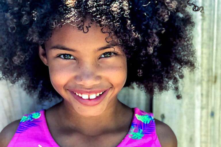 Photo d'une petite fille métisse aux cheveux frisés qui souris face à l'objectif.
